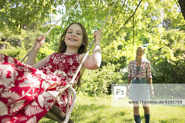 Summer. A Girl In A Sundress On A Swing Under A Leafy Tree. A Woman Standing Behind Her.