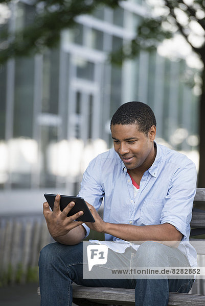 Summer. A Man Sitting On A Bench Using A Digital Tablet.