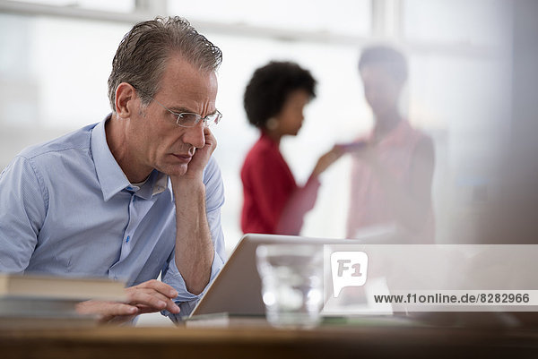 Büroleben. Ein Mann sitzt an einem Computer-Laptop  auf einen Arm gelehnt  und zwei Frauen im Hintergrund.