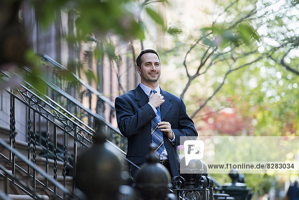 Ein Mann im Geschäftsanzug beim Anpassen seiner Krawatte. Am Fuße der Stufen eines Stadthauses in einer Terrasse.