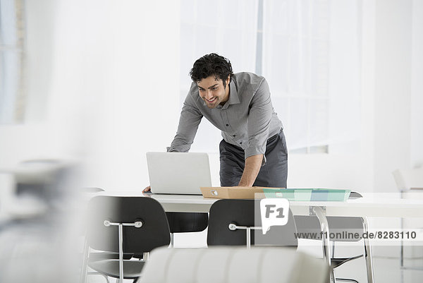 Business. A Man Standing Over A Desk  Leaning Down To Use A Laptop Computer.