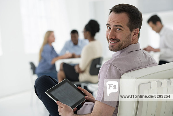 Office Interior. Meeting. One Person Looking Over His Shoulder And Away From The Group. Holding A Digital Tablet.