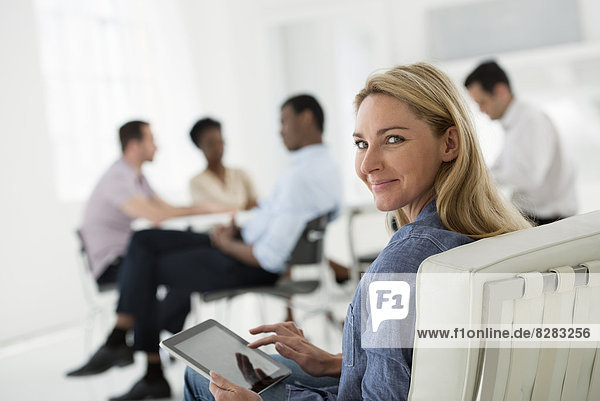 Office Interior. Meeting. One Person Looking Over Her Shoulder And Away From The Group. Holding A Digital Tablet.