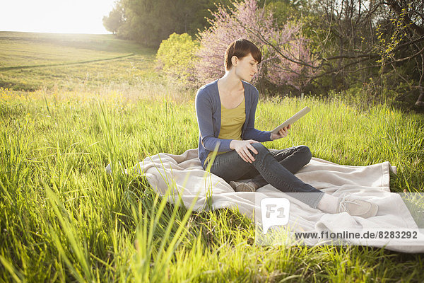 Eine junge Frau sitzt auf einem Feld  auf einer Decke  hält ein digitales Tablett in der Hand und schaut auf den Bildschirm. Arbeit im Freien.
