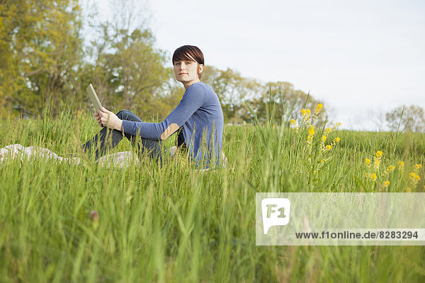 A Young Woman Sitting In An Open Space  A Grass Field  On A Blanket  Holding A Digital Tablet.