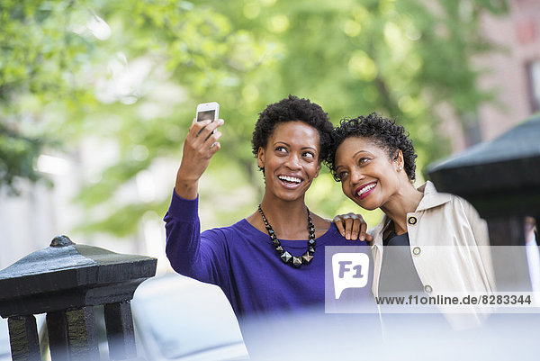 Stadtleben. Zwei Frauen sitzen auf einer Parkbank  Seite an Seite. Posieren für ein Foto mit einem Smartphone.