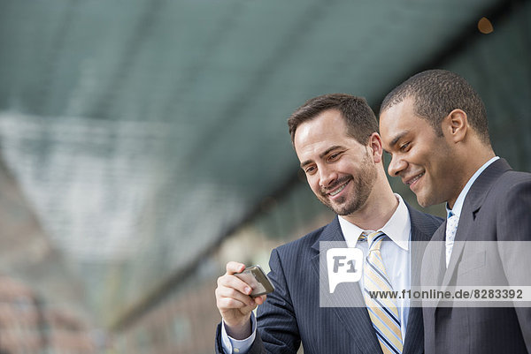 Stadt. Zwei Männer in Geschäftsanzügen  die lächelnd auf ein Smartphone schauen.