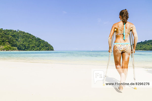 Thailand  Koh Surin island  woman with crutches walking at the white sandy beach