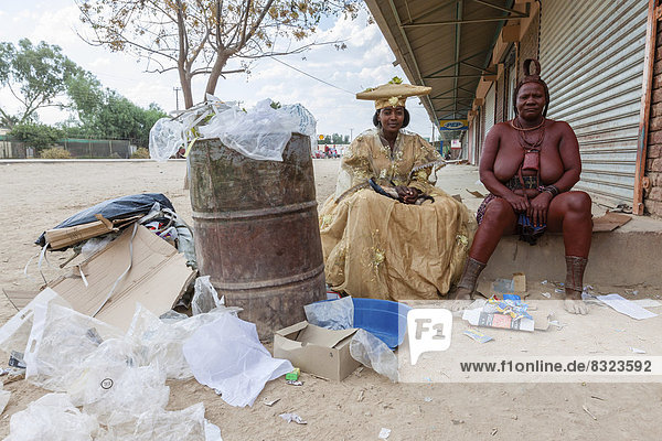 Herero-Frau und Himba-Frau in traditioneller Kleidung sitzen neben Müll