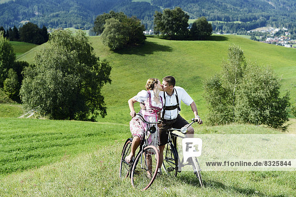 Man wearing leather pants and a woman wearing a dirndl kissing on old bicycles within a natural landscape