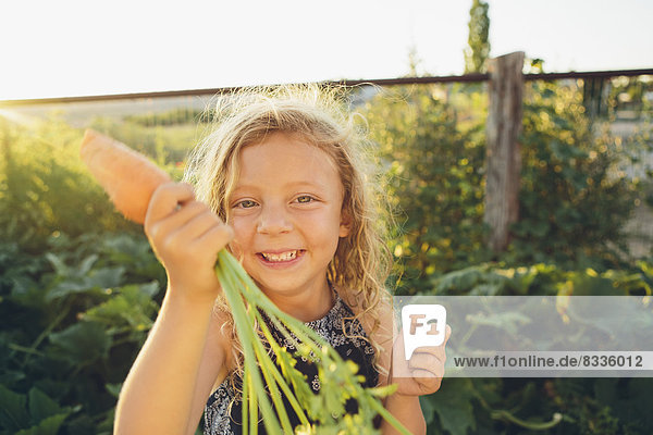Ein junges Mädchen mit langen roten Lockenhaaren im Freien in einem Garten mit einer großen frisch gepflückten Karotte in der Hand.