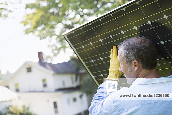 Ein Mann  der ein Solarpanel zu einem im Bau befindlichen Gebäude trägt.