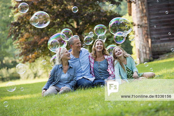 Eine Familie sitzt auf dem Rasen vor einer Bar  bläst Seifenblasen und lacht.