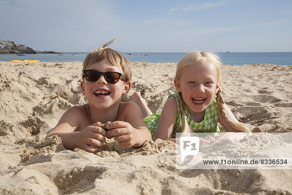 Ein Junge und ein Mädchen liegen mit dem Bauch auf dem Sand  lachend und in die Kamera blickend.
