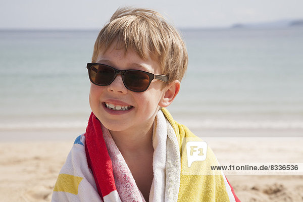 A boy in sunglasses on the beach  with a towel around his shoulders.