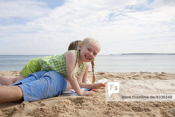 Ein Junge liegt vorne auf dem Sand und schaut aufs Meer hinaus. Seine Schwester liegt auf ihm.