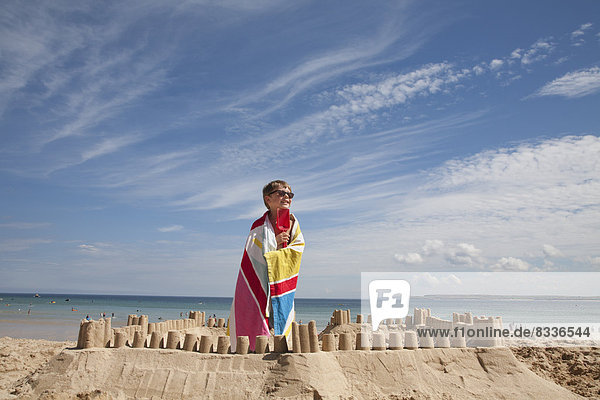 A boy standing beside a sandcastle  on top of a mound of sand. Beach.