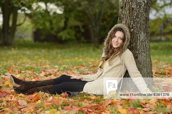 Lächelnde junge Frau sitzt unter einem Baum im Herbst