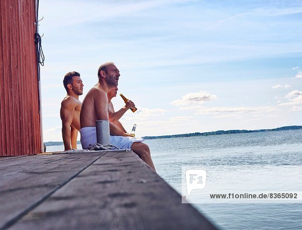 Drei männliche Freunde sitzen vor der Sauna und genießen Bier.