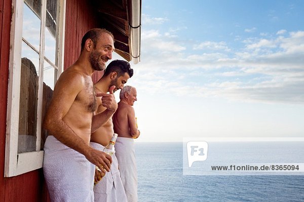 Drei männliche Freunde stehen vor der Sauna und genießen Bier.