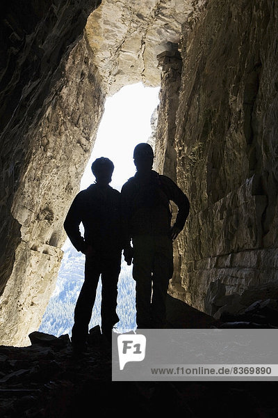 Silhouette Of Two Male Hikers In Cave With Entrance Glowing In The Background Bragg Creek  Alberta  Canada