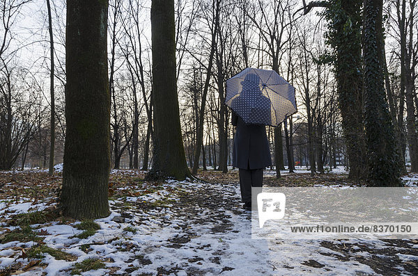 A Woman Holding An Umbrella And Standing In A Park Area Surrounded By Trees With A Trace Of Snow