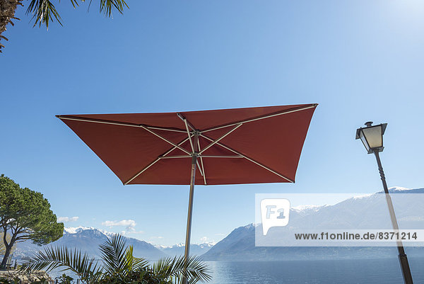 A Patio Umbrella And Lamp Post Against A Blue Sky With A View Of Lake Maggiore And The Swiss Alps Brissago  Ticino  Switzerland