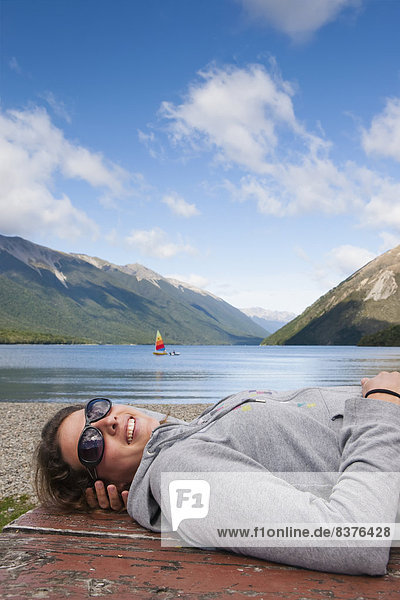 A Girl Lies Down On A Picnic Table And Enjoys The Sun With A Sailboat In Lake Rotoiti In The Background  Tasman Region  New Zealand