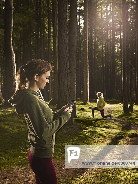 Frau steht im Wald und schaut auf ihr Smartphone  hinten macht eine andere Frau Dehnübungen  Tirol  Austria