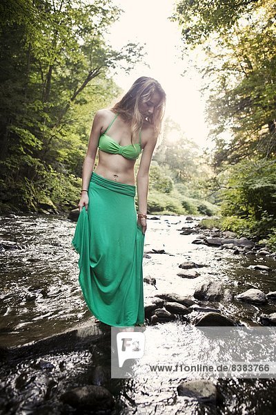 A young woman wearing a bikini dips her toes in a woodland stream.