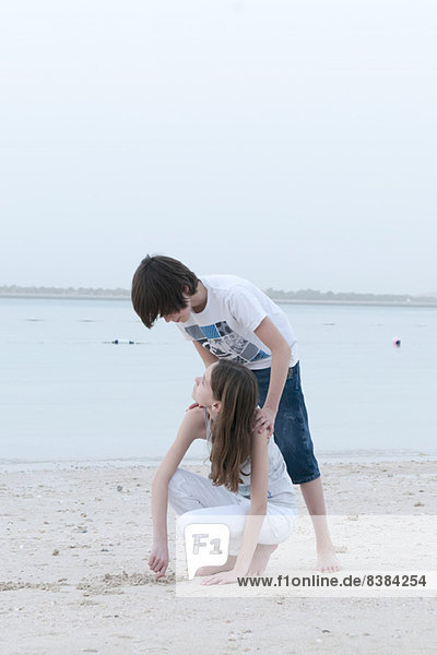 Teenager-Junge schaut der Schwester über die Schulter  als sie am Strand im Sand gräbt.