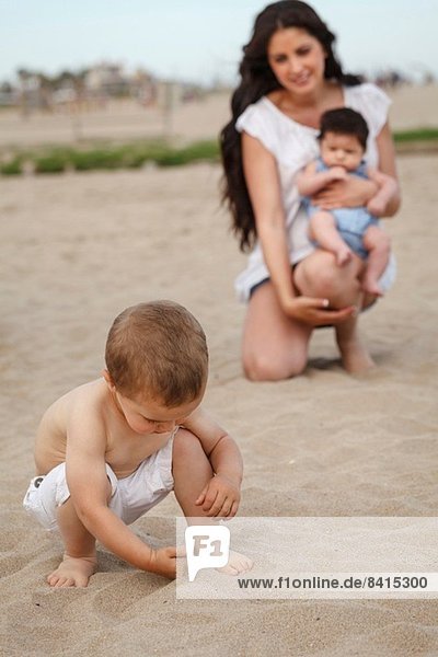 Toddler digging in sand with hand  mother and baby in background