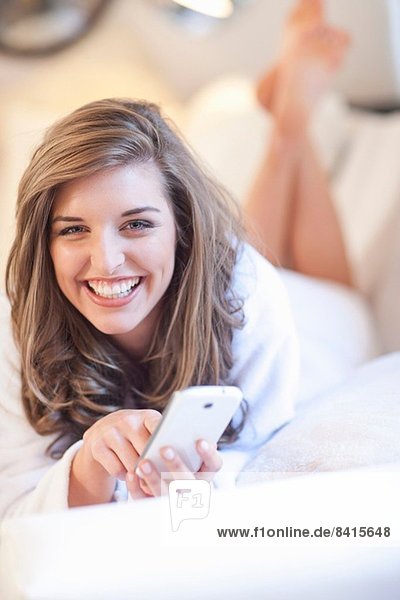 Porträt einer jungen Frau auf dem Bett liegend mit Handy