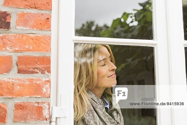 Mittlere erwachsene Frau am Fenster mit geschlossenen Augen  lächelnd