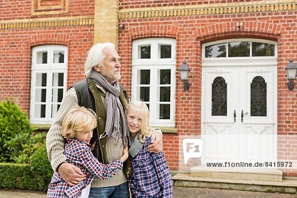 Großvater mit Armen um Enkelkinder vor dem Haus