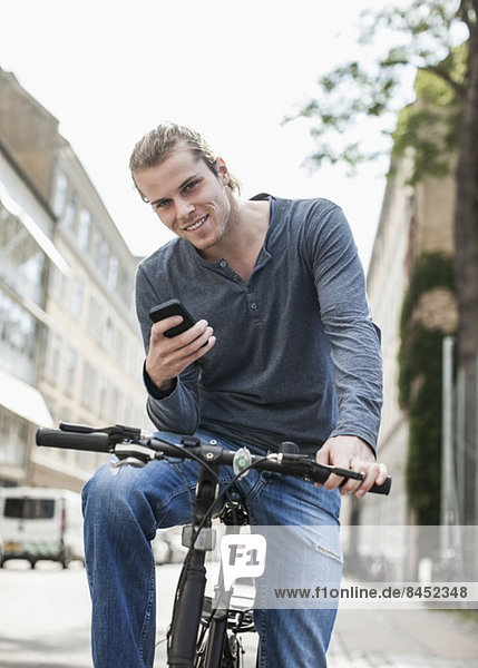 Portrait of confident young man with mobile phone sitting on bicycle on street