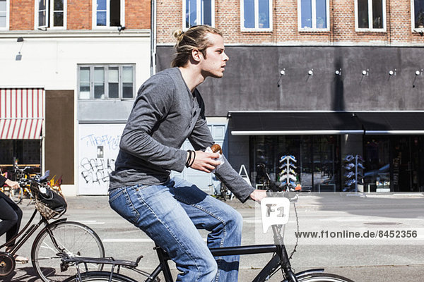 Profilaufnahme eines jungen Mannes  der Burger hält  während er Fahrrad fährt.