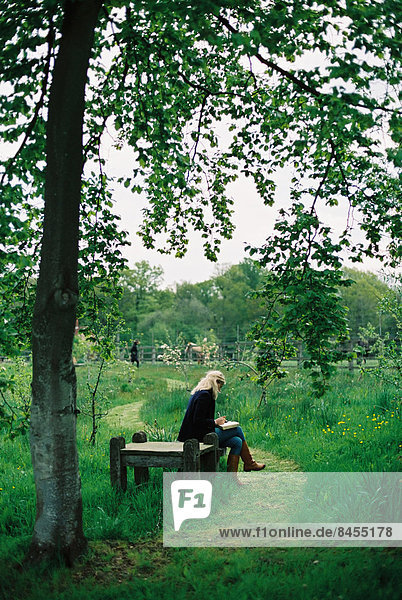 Trees and long grass. A mown path. A woman seated on a wooden seat.