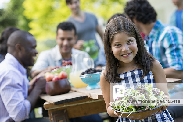 Adults and children around a table in a garden. A child holding a bowl of salad.