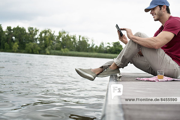 Ein Mann sitzt auf einem Steg an einem See und benutzt ein digitales Tablett.
