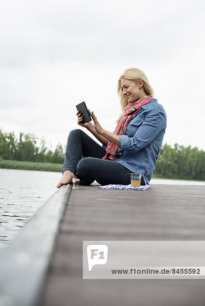 A woman sitting on a jetty by a lake  using a digital tablet.