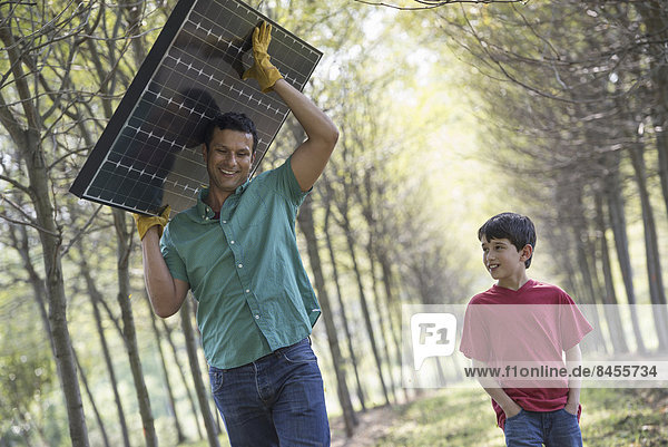 A man carrying a solar panel down an avenue of trees  accompanied by a child.