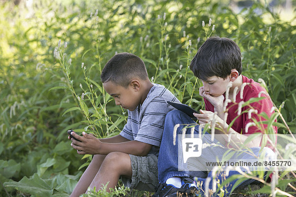 Two boys sitting in a field  one on a smart phone and one using a digital tablet.
