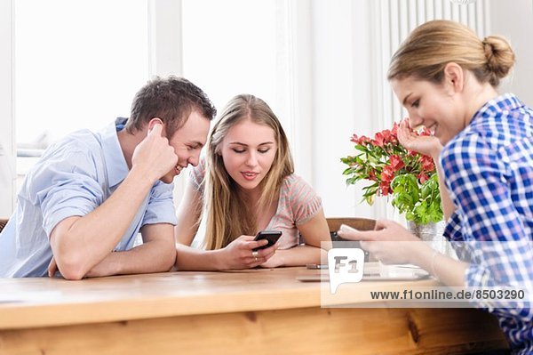Drei junge Leute sitzen an einem Tisch mit dem Handy und lächeln