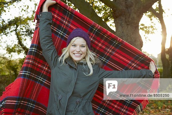 Porträt einer jungen Frau im Park  die eine Tartan-Picknickdecke hochhält.