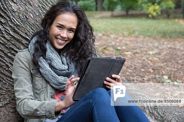 Junge Frau die sich mit einem digitalen Tablett an den Baum lehnt