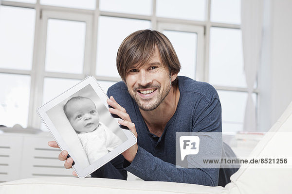 Mann auf Sofa liegend  mit digitalem Tablett und Foto des Neugeborenen
