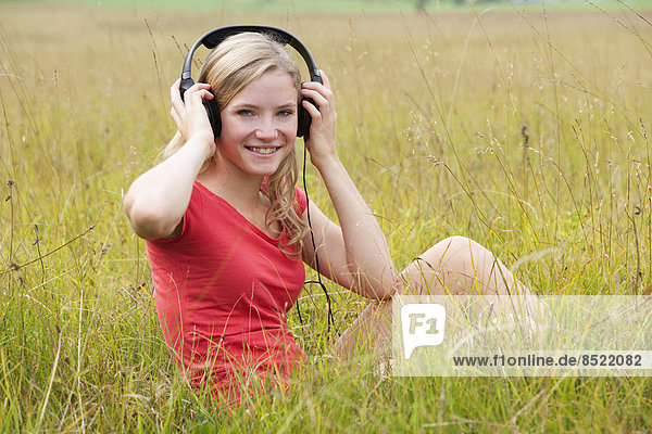 Österreich  Salzkammergut  Mondsee  junge lächelnde Frau mit Kopfhörer auf einer Wiese sitzend