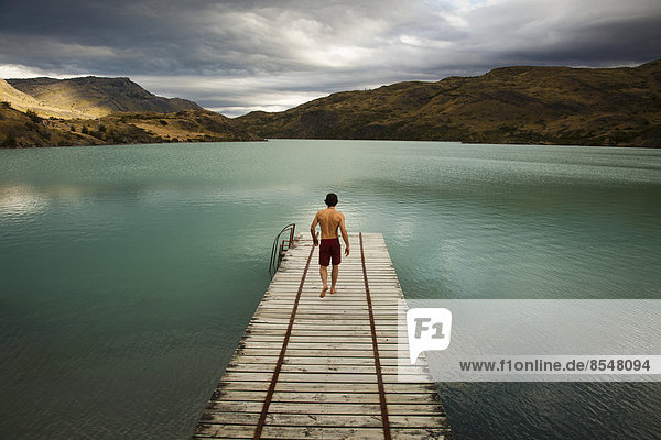 A young man walking down a wooden pier  towards calm lake surrounded by mountains in Torres del Paine National Park  Chile.