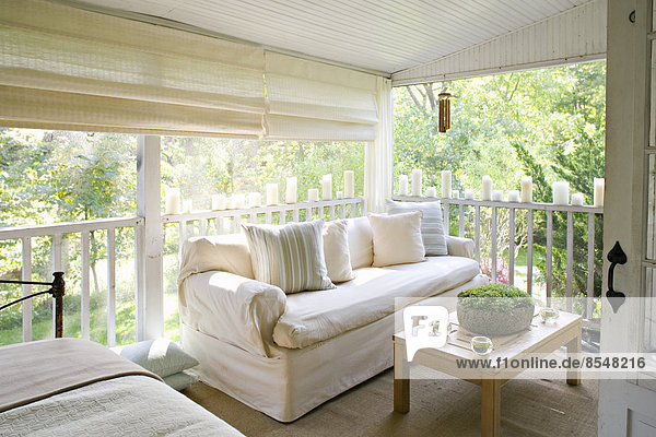 A veranda or shaded porch of a house in the woods  with cream sofa and candles along the balustrade.